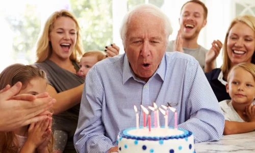 Elderly Man Blowing Out Birthday Candles With Family Around Him
